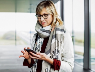 Blonde Caucasian woman with a sweater, scarf and glasses on looking down at her cell phone.
