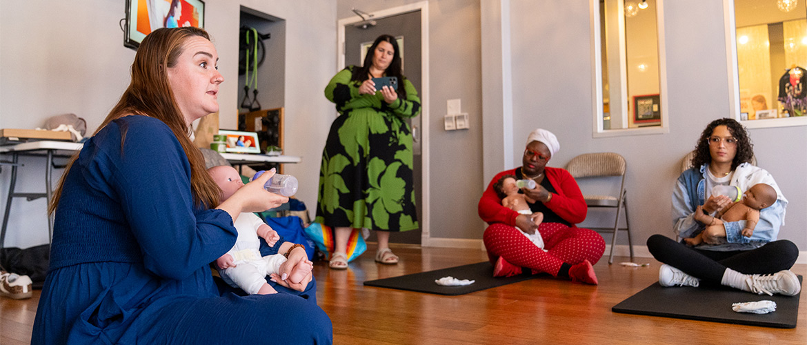 Women holding training baby dolls and bottles sit on mats during a doula workshop.