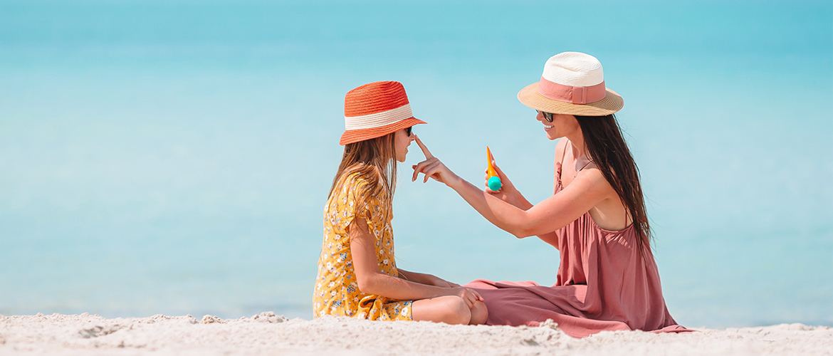 A woman holding sunscreen touches a child's nose as they sit on the beach