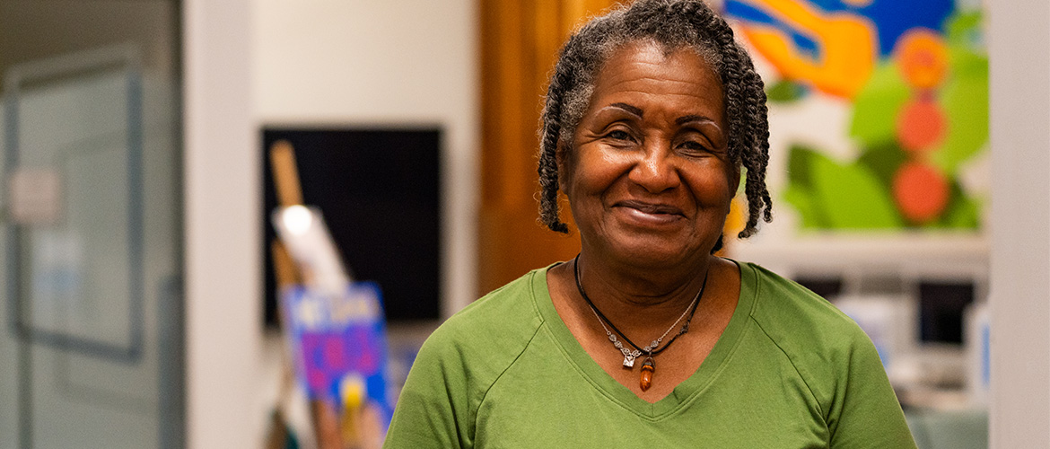 An older woman wearing a green shirt smiles in a room with brightly colored art in the background 