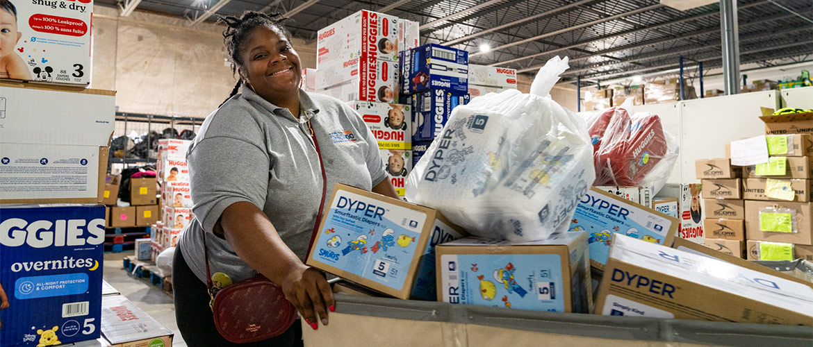 A smiling woman in a warehouse fills a large cart with boxes of diapers