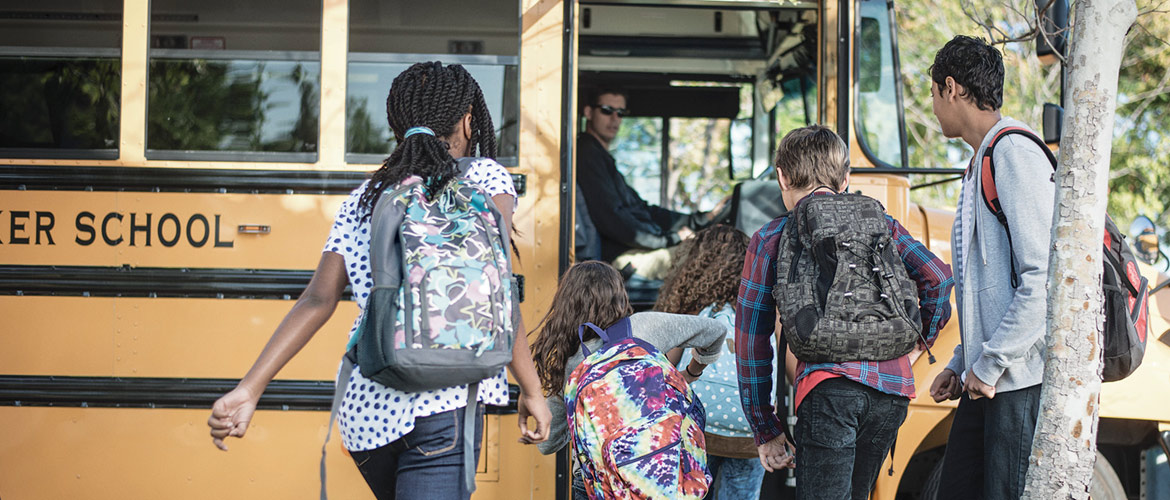 Five children are greeted by a driver as they board a yellow school bus