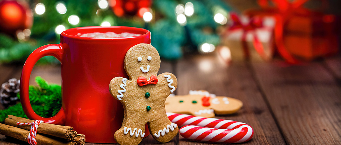 A gingerbread man propped against a red mug of hot chocolate next to a small bunch of cinnamon sticks 