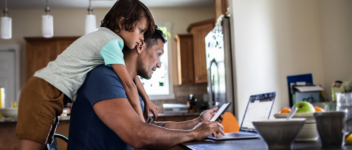 A child hangs over a man who is looking at his cell phone and working on his laptop.