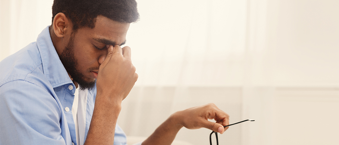 Man sits with his eyes closed, pinching the bridge of his nose with one hand while holding a pair of glasses in his other hand