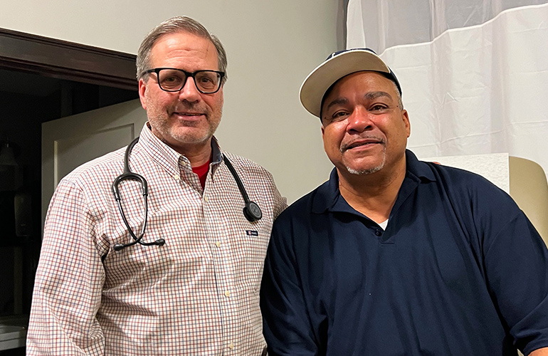 Middle age man in glasses and stethescope around his neck stands with man in blue shirt and hat