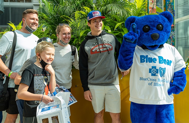 A NASCAR driver and BCBSIL mascot Blue Bear pose with a family in a hospital atrium. 