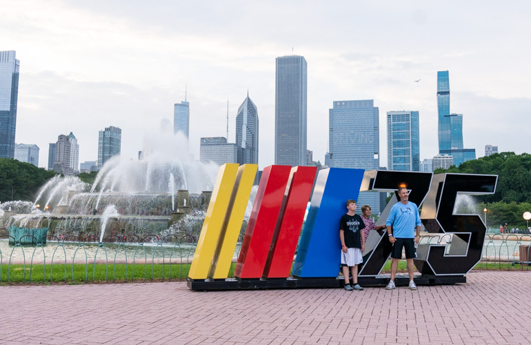 Two children and a man stand with a large yellow, red and blue sculpture with the number 75 in black and Buckingham Fountain in the background. 