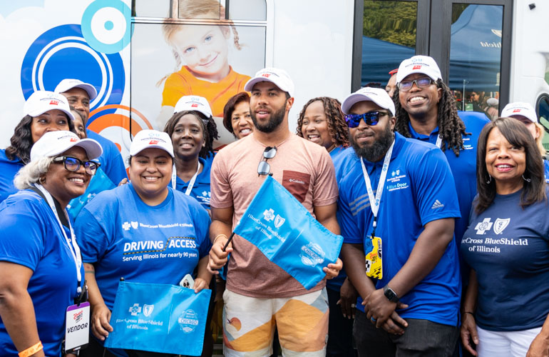 NASCAR driver Bubba Wallace wearing a baseball cap and shorts stands with a group of BCBSIL employees wearing blue shirts with the BCBSIL logo. 