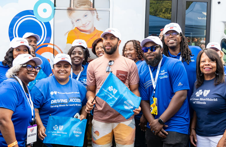 NASCAR driver Bubba Wallace wearing a baseball cap and shorts stands with a group of BCBSIL employees wearing blue shirts with the BCBSIL logo. 