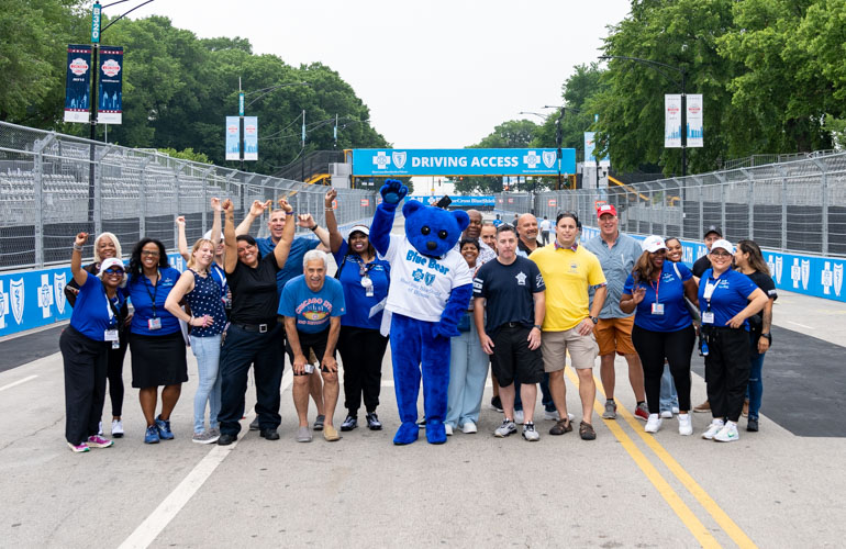 A group of people pose with BCBSIL mascot Blue Bear with the words Driving Access seen in the background in large letters on a bridge over the racecourse. 