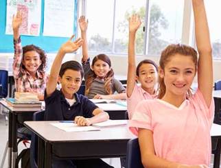 Niños en edad de escuela media sentados en sus pupitres en un aula con las manos levantadas.