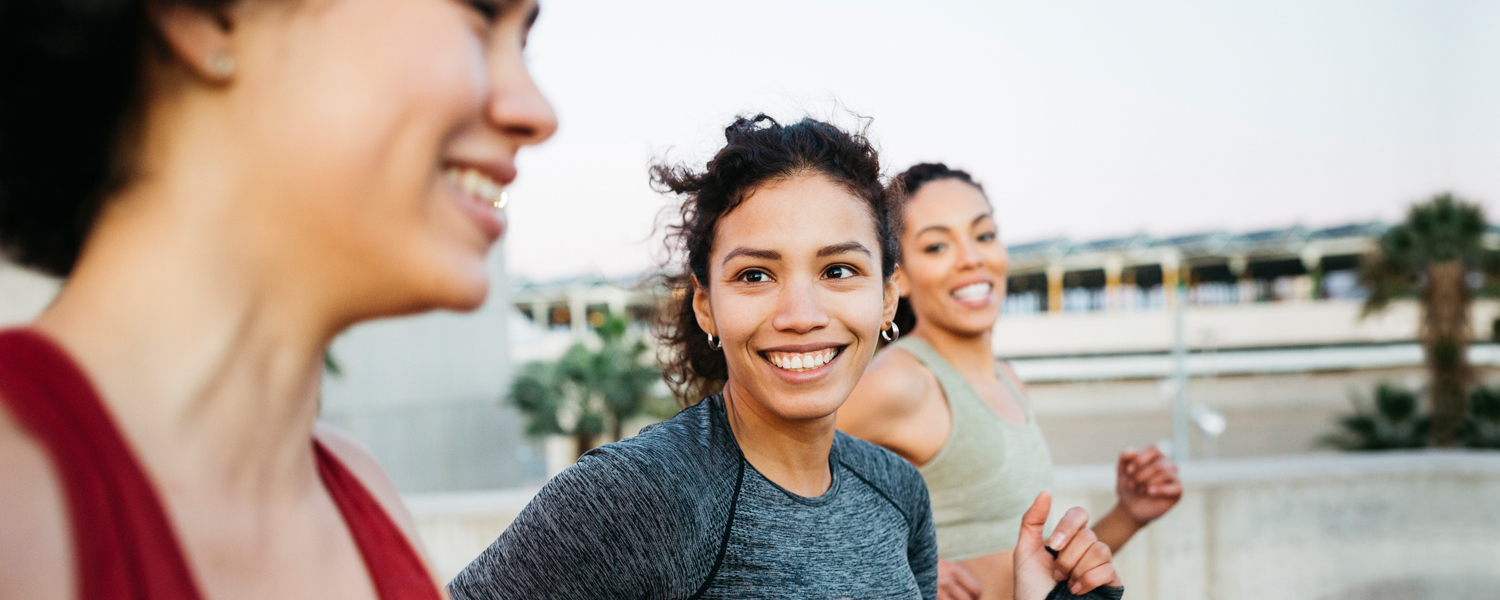 A woman jogging outdoors smiles widely at another woman in the foreground