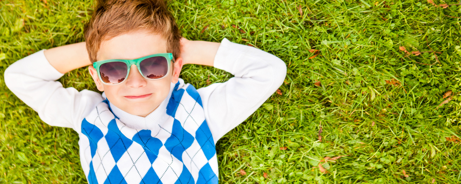 A young boy wearing sunglasses lies in the grass with his hands behind his head
