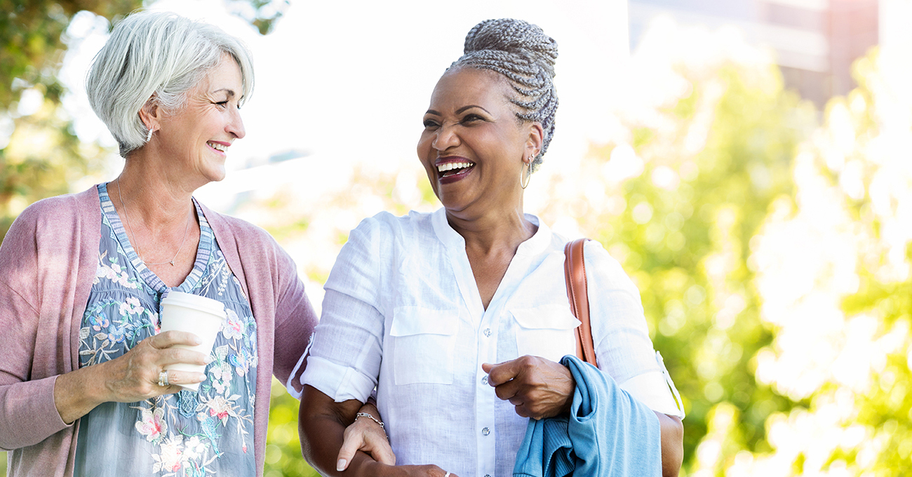 Joyful senior women laughing as they walk arm in arm on a sunny day. One woman is holding a paper coffee cup. 
